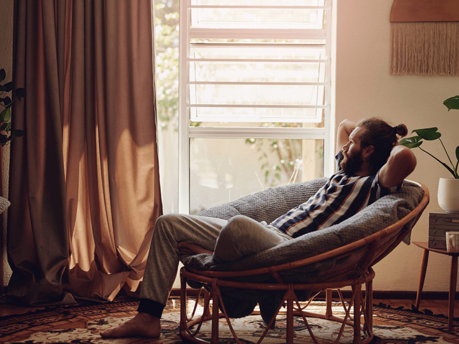 Man reclines in a shell shaped chair while looking out the window of his home.