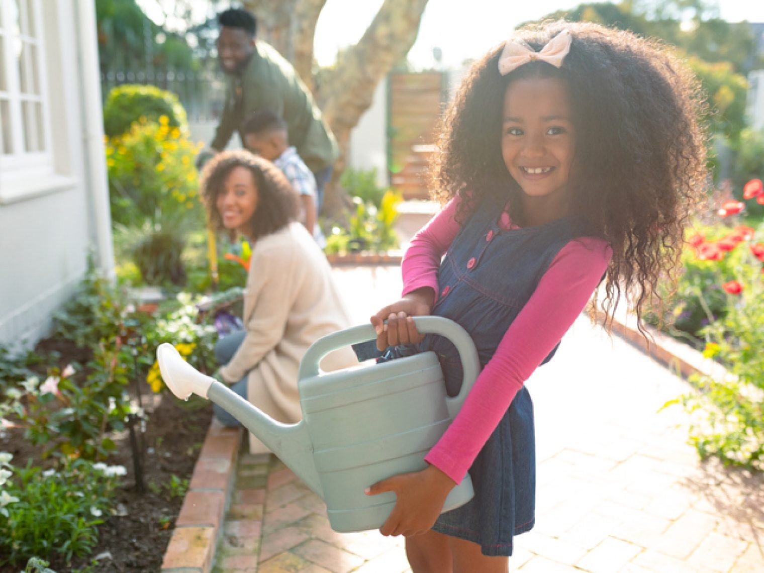 A young girl holding a watering can smiles at the camera, with her family gardening together in the background, creating a heartwarming scene of outdoor family activity.