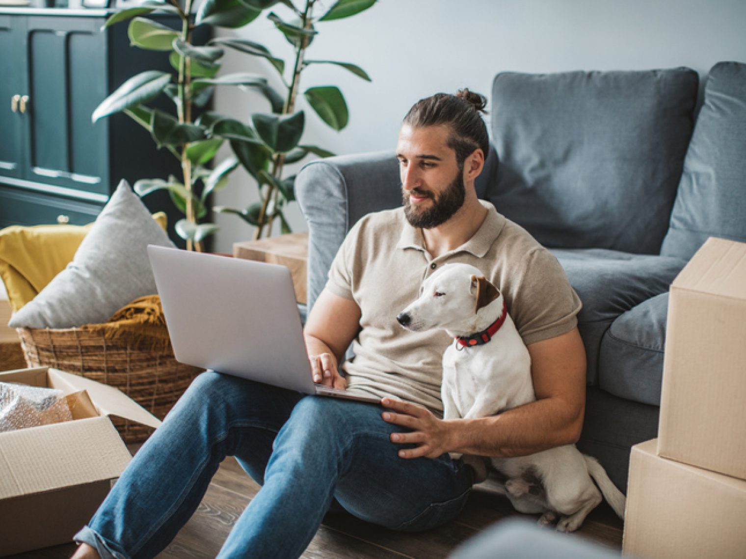 A man with a laptop sits comfortably on the floor against a sofa, accompanied by his dog, in a living room with moving boxes.