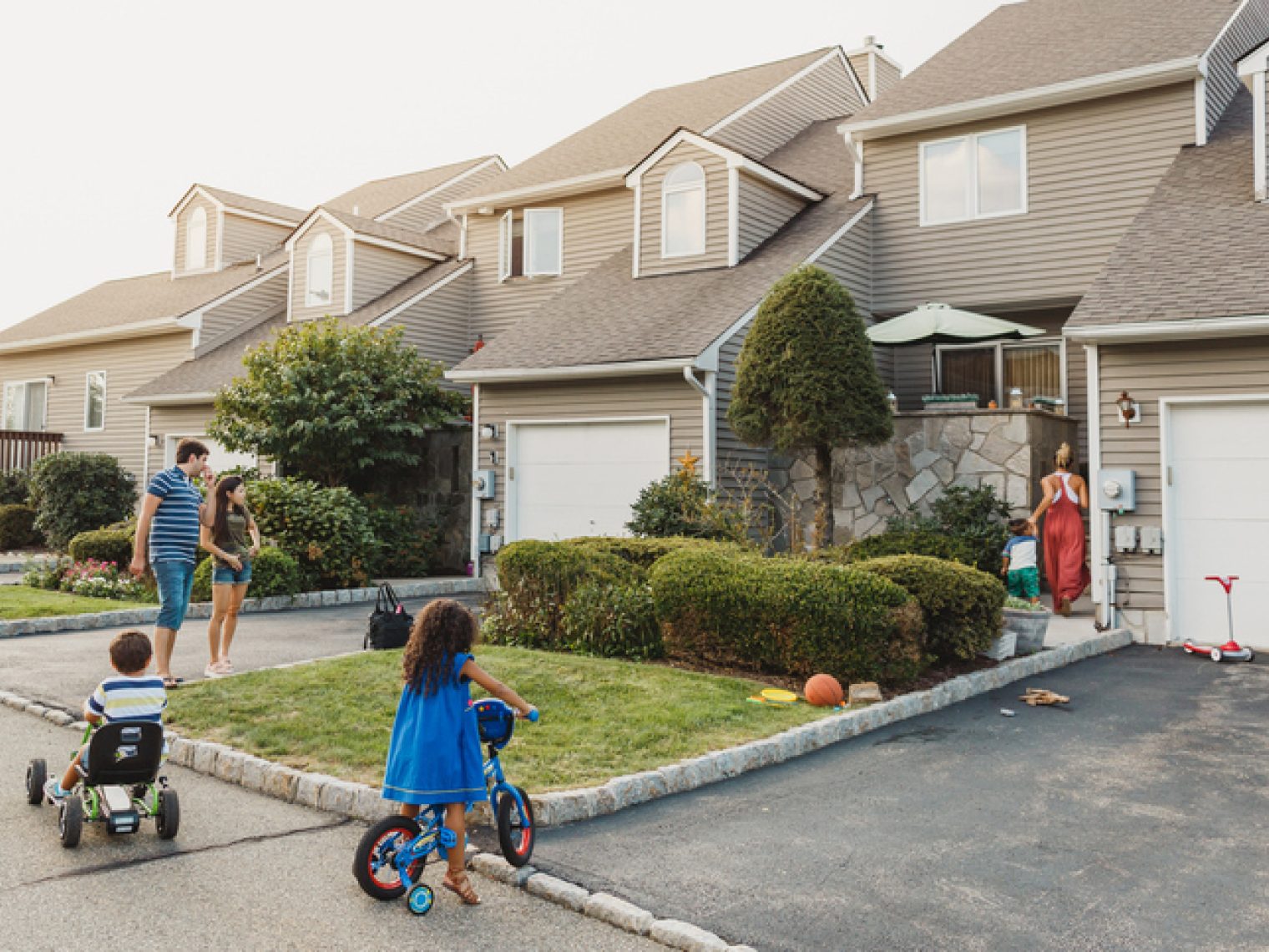 A family is enjoying a sunny day outside their suburban home; two children are playing with their bikes on the driveway, while the adults are in the background by the garage.