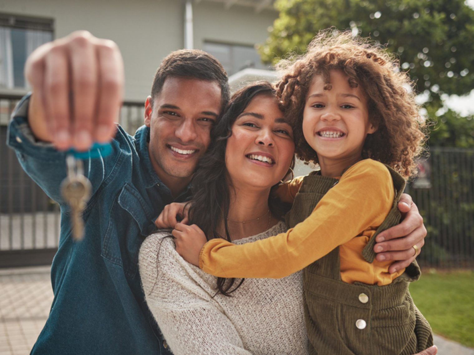 A joyful family with a young child smiles at the camera, with one of the parents holding up a set of keys, suggesting they have just bought a new home.