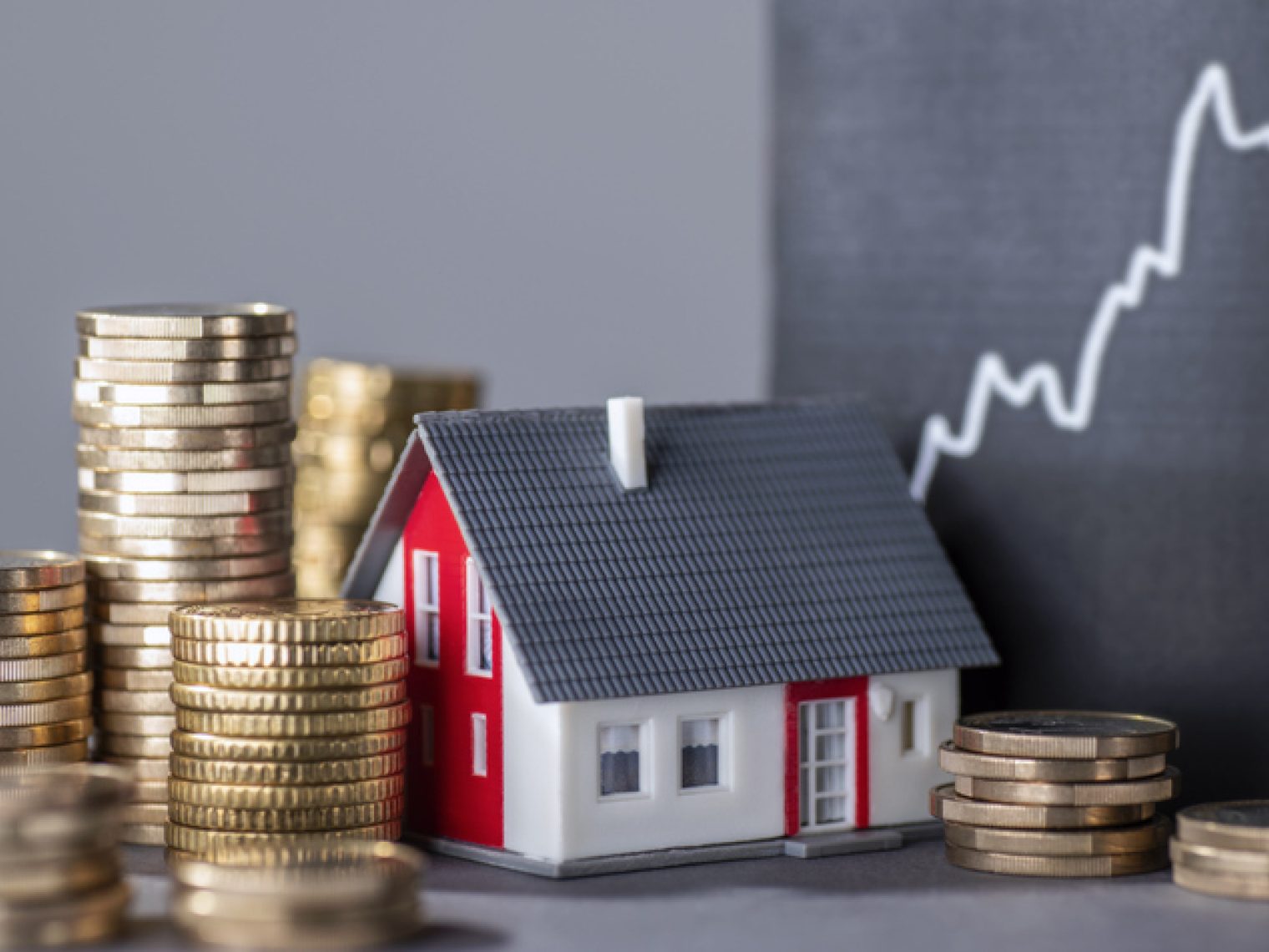 A small red and white model house is surrounded by stacks of coins with a rising chart in the background.