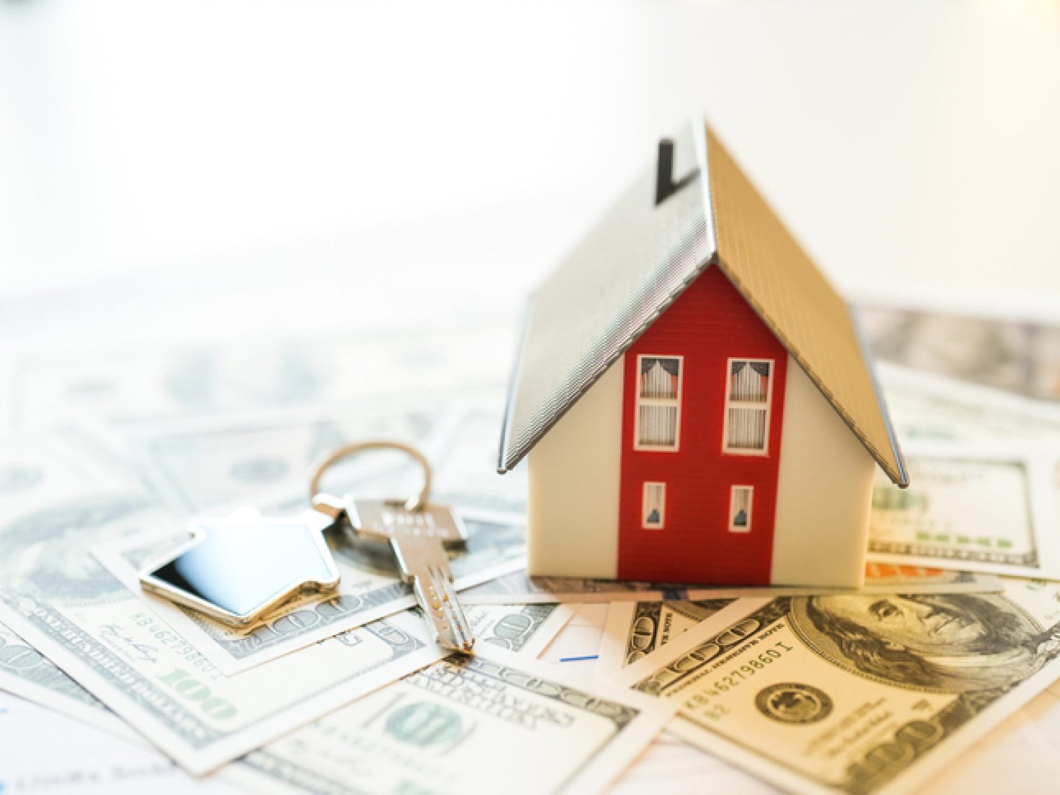 Miniature house on dollars on table with a pair of house keys to the side.