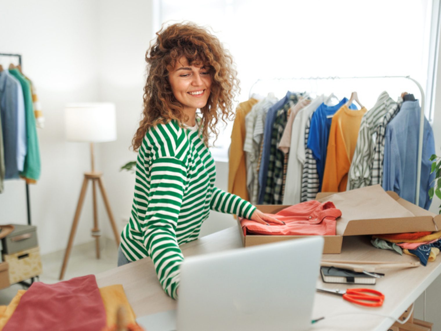 A cheerful woman with curly hair, wearing a striped shirt, is packaging clothes in a box, with a laptop open in front of her.