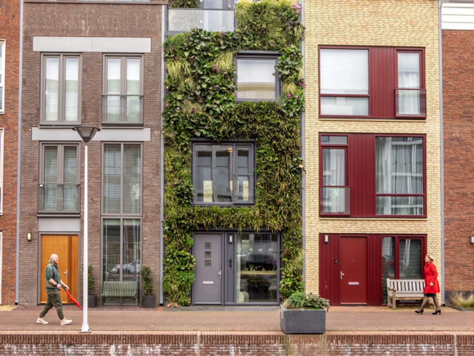 A street view of modern townhouses, one with a vertical garden facade, as pedestrians walk by on the sidewalk, offering a glimpse into urban residential architecture.