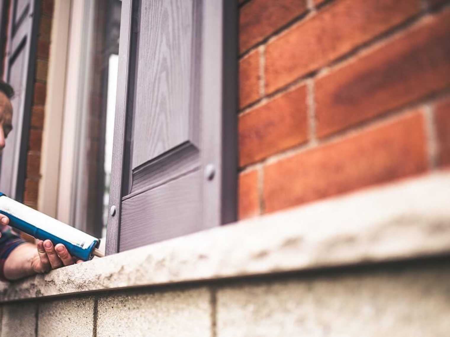 A man applies caulk to the window of a brick home.