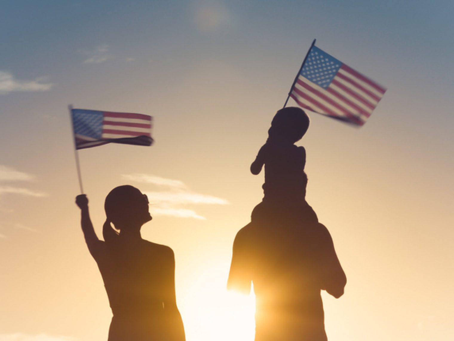 A silhouette of a family with an adult holding an American flag and a child on someone's shoulders also waving a smaller flag, against a sunset sky.