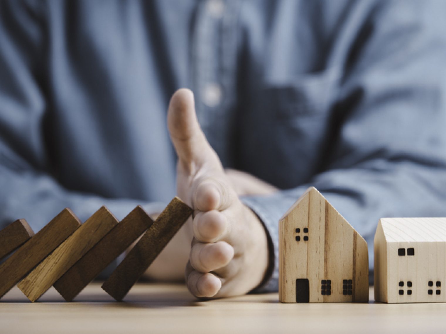 A person's hand stopping falling wooden dominoes before they reach a model house.