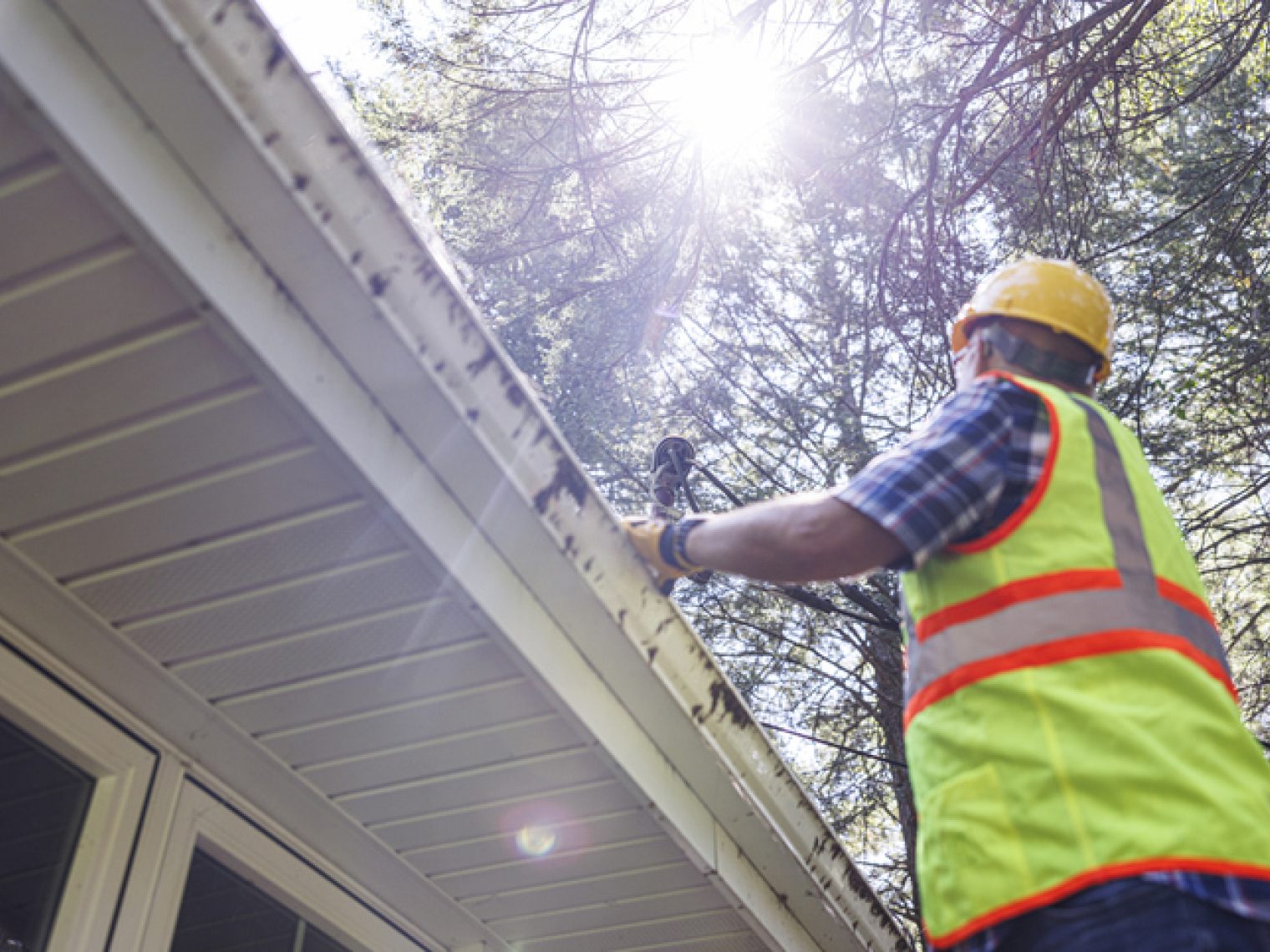 A worker in a high-visibility vest and hard hat repairs or inspects a house's gutter, with sunlight filtering through the trees overhead.