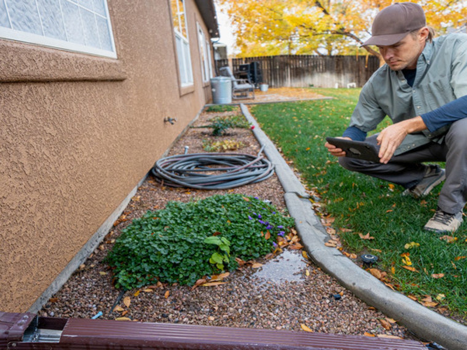 A man in work clothes and a cap is kneeling with a tablet in a backyard, surrounded by plants, a coiled hose, and fallen leaves, possibly engaged in landscaping or maintenance.