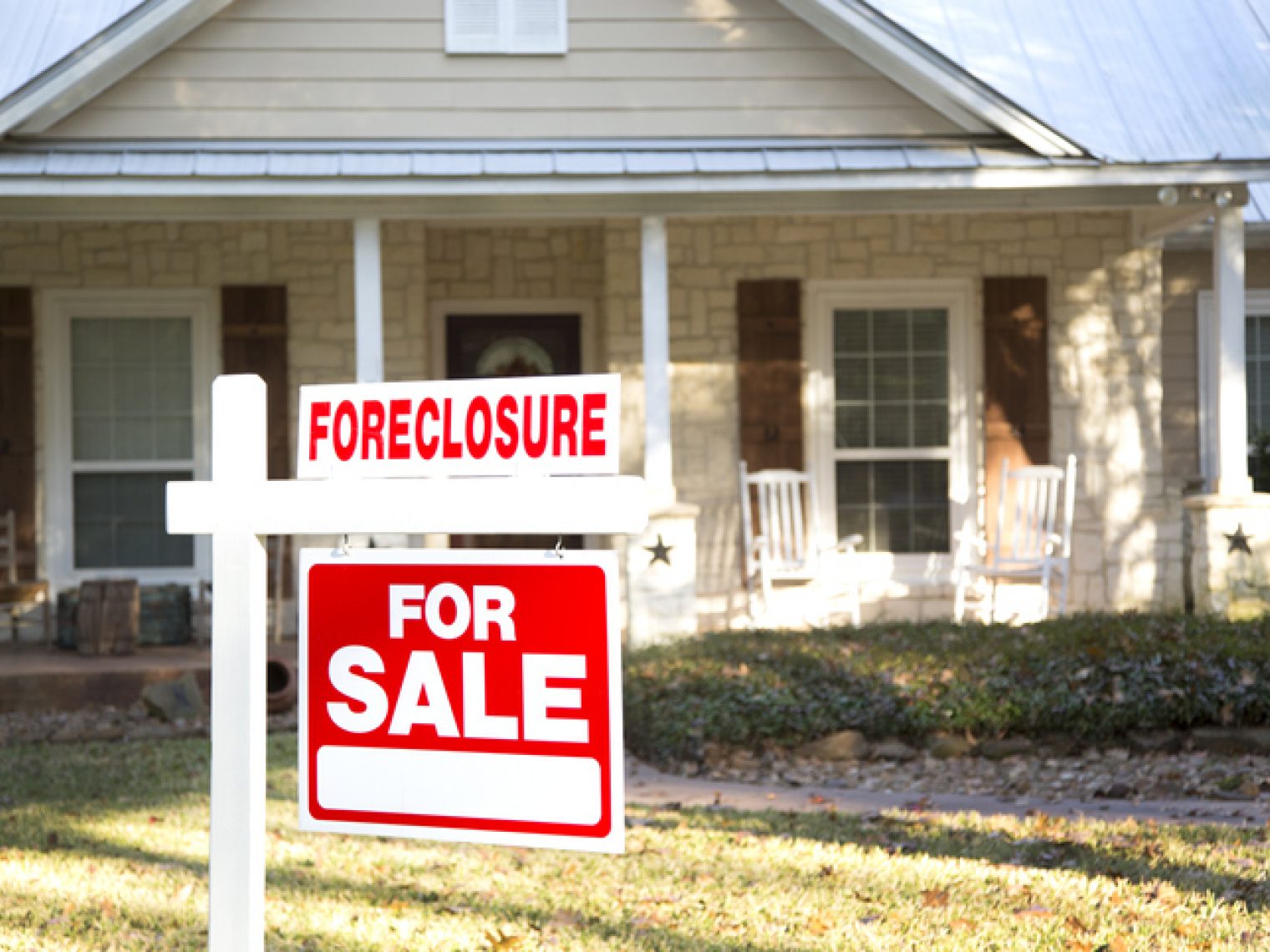 A "For Sale" sign with an additional "Foreclosure" rider is prominently displayed in the front yard of a single-story house, indicating that the property is on the market due to foreclosure.