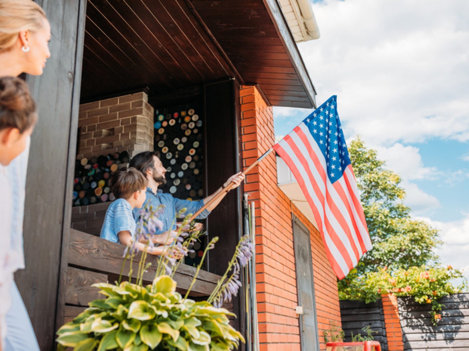 A family is engaged in a patriotic moment, with a man and child holding the American flag out from the porch of their home, as others watch with pride.