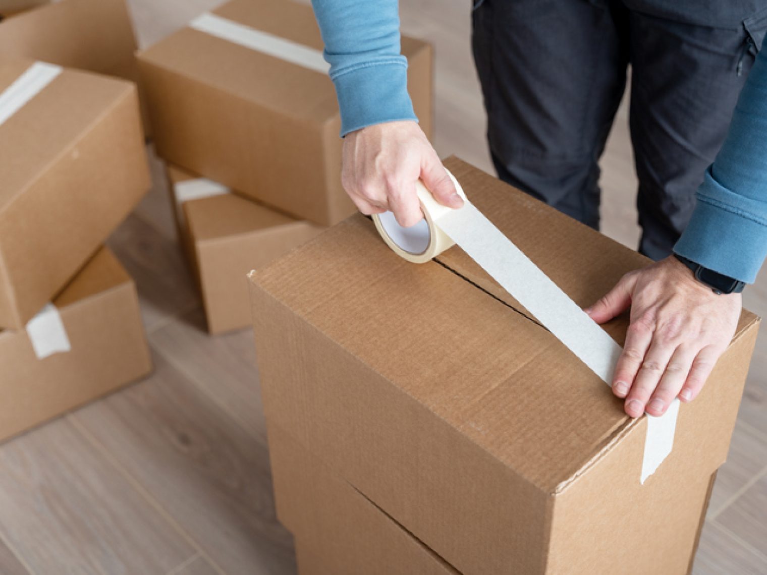 A person is taping a cardboard box shut, part of a stack of boxes, indicating packing or moving activities.