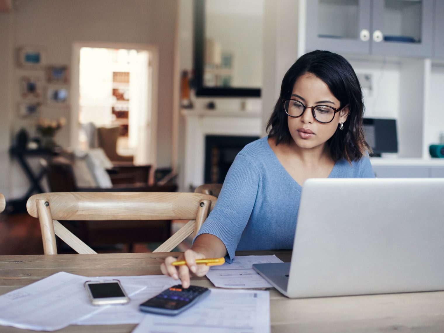 Focused woman wearing glasses and a blue sweater working at a wooden table with a laptop, calculator, papers, and a smartphone, in a cozy home setting.